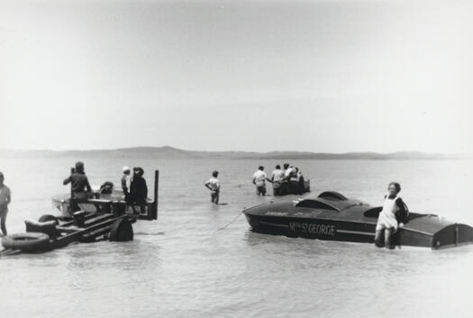 Lake George Sailing Club regatta. Photo shows a female leaning against a motor boat, a group of 5 men a little futher out on the lake, and 4 men closer to the shore on a boat trailer.