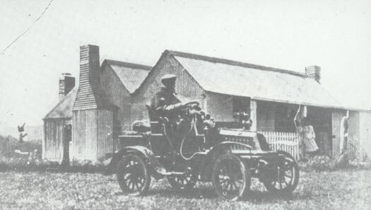 Man sitting in a car outside a house at Round Hill. A woman can be seen on the verandah.