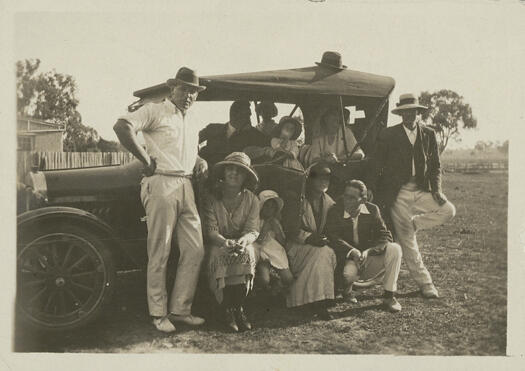 Ted Shumack (at left) and a group of people with a car, at the Ainslie Cricket Ground, Yass Road. A.W. (Eddie) Edwards is kneeling and Fred Cox is standing at right. All three men are dressed in cricket whites. Part of an old building can be seen in the background.