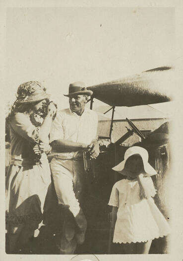 Ted Shumack talking to Minnie Pooley with a small girl standing next to a car. Shumack is dressed in cricket whites.