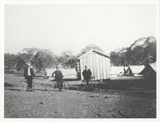 Surveyors camp on the slopes of Capital (Kurrajong) Hill. Shows three men with push bikes standing in front of a building with tents in the background.