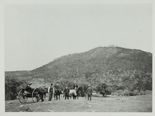 Photo shows a man with a white beard sitting in a sulky, two men in horse back with four men standing. Black Mountain is in the background.