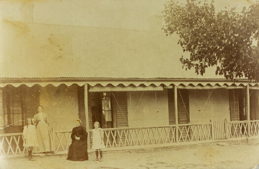 Elderly woman sitting in a chair in front of a verandah with a small girl standing beside her. There is another woman standing on the verandah with a small girl.
