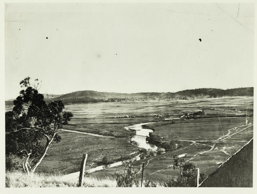 The Power House is shown on the right hand side with a plume of steam. In the foreground a railway bridge crosses the Molonglo River.