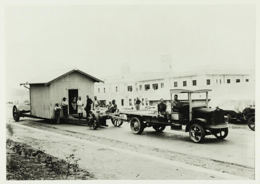 Truck towing a trailer removing a storage hut used in the construction of Parliament House. Eight men are looking on.