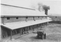 Canberra Brickworks at Yarralumla, showing the front of the ovens, smoke coming from the chimney and a man standing next to a car in the yard in front of the kilns.