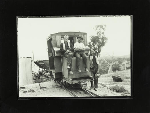 Rail car pulling two hoppers with two men on the engine and one man leaning against the engine.
