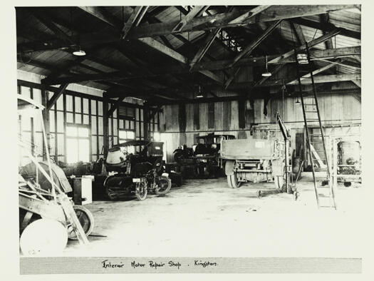Photo of the interior of a motor repair shop at Kingston showing a truck, motor bike and side car and two cars, with one on a hoist. 