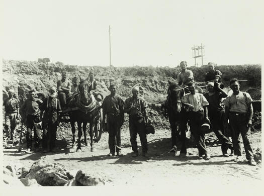 Construction workers building an unidentified road with two horses and carts.