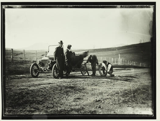 Two men changing the back left hand side tyre being watched by two other men. The vehicle is jacked-up. The scenery is wide open with no identifying features.