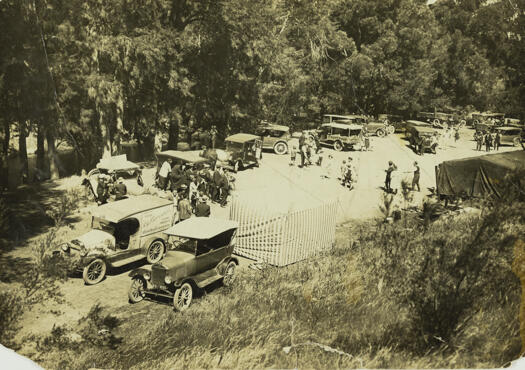 About 16 vehicles near some trees wityh a couple of tents in the foreground. Men in suits and woman and children are present.