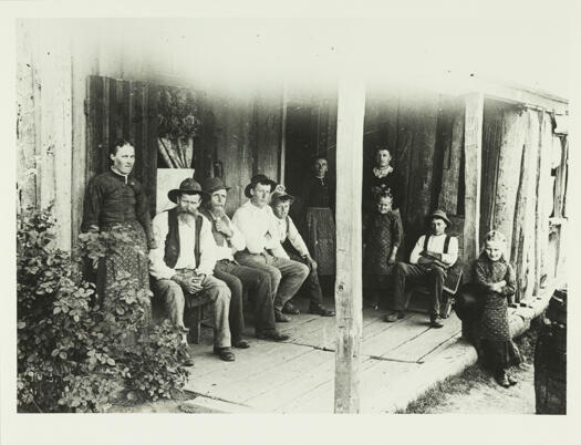 A family group on the verandah of a slab hut. Shows five males and five females.