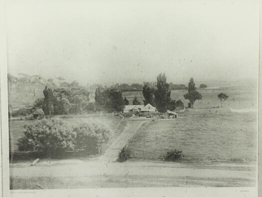 Shows a road in the near foreground with a driveway into an unknown farmhouse in the middle distance. The farmhouse is surrounded by trees.