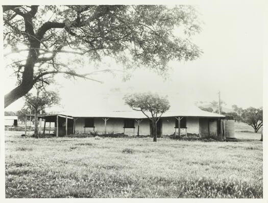 A front view of Rose Cottage. A water tank can be seen on the right side of the cottage. On the left side is a partial view of a shearing shed and sheep pens.