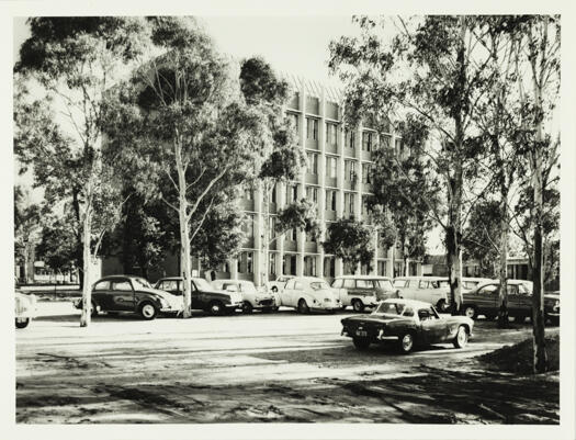 ANU Chancellery visible through trees. A large number of cars are parked on the street.