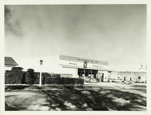 A side view of W.B. Freebody's garage. Petrol bowsers and parked cars shown to the right of the picture.