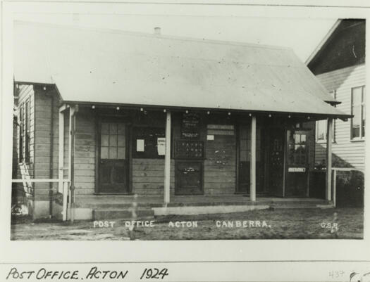 Front view of the Post Office, Acton. It shows a verandah with a notice board, approximately 18 post boxes and a telephone box on the right hand side.