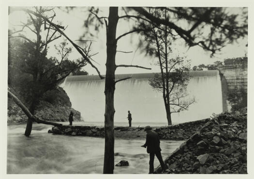 Cotter Dam wall with water flowing over the top. Two men are seen fishing in the river below the dam.