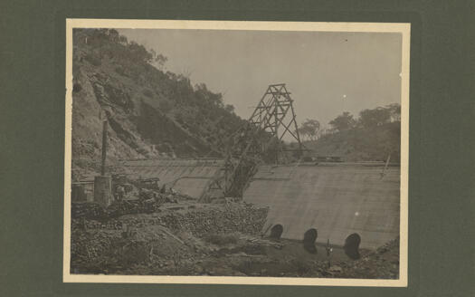 Cotter Dam under construction, showing the base of the dam, from below the dam site. There is a frame mounted on the front of the wall so that concrete can be pumped to the top of the wall.