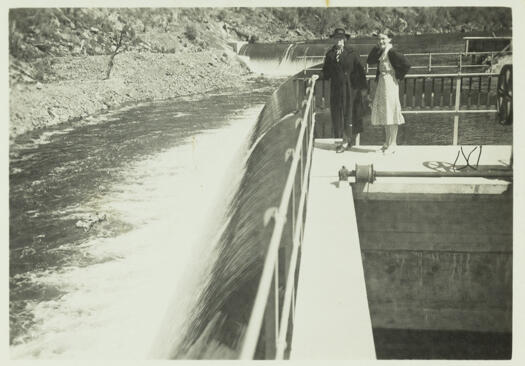 Two women standing on the walkway of the Cotter Dam with water flowing over the wall.