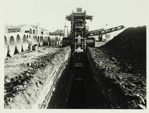Shows a ditcher machine digging a trench with concrete pipes on one side and a heap of dirt on the other side of the trench.