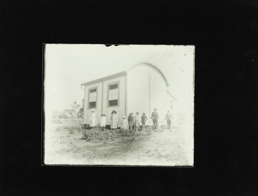 Early Canberra school with nine children standing outside. Two of the boys have bicycles.