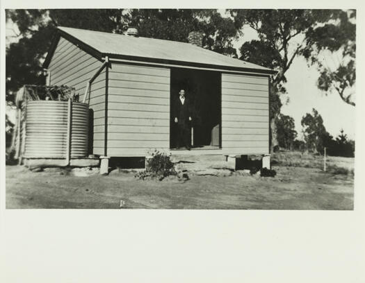 Mulligans Flat School (since removed), showing a small timber building with steps to the entrance. On the right side is a water tank. The teacher, James Gormly, is standing in the entrance wearing a suit and bow tie.