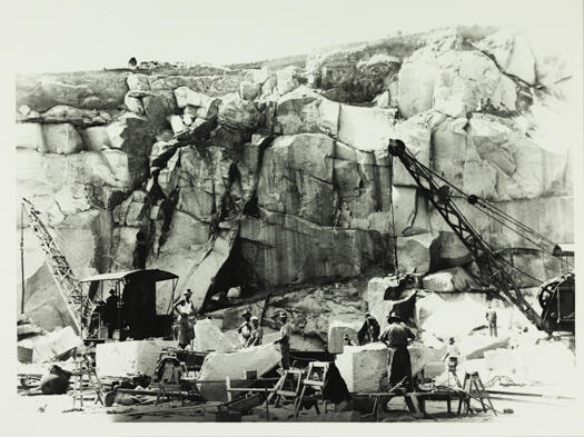 Shows a quarry face with rocks cut to size and men working on them. Two cranes are in operation.