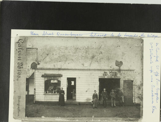 Shops in Crawford Street, Queanbeyan, opposite the Fire Brigade. Two shops are shown; Fancy Goods Shop, with a woman standing in front and Pike's Hay & Corn shop, with three men and boy standing in front.