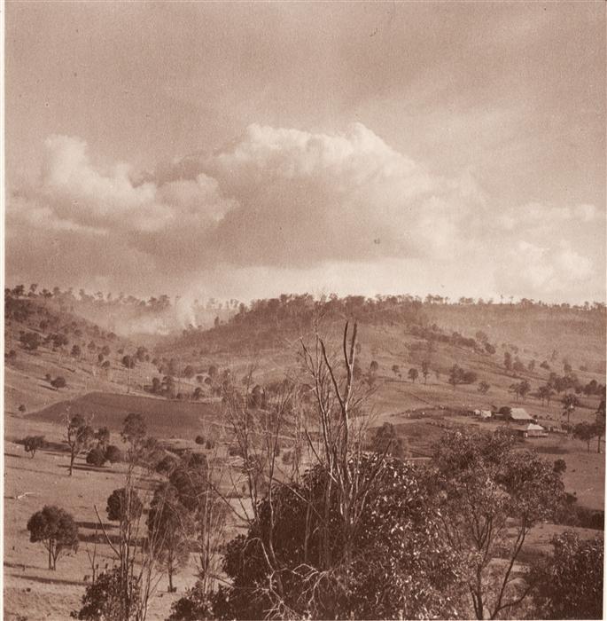 Bushfire burning over a hill. An unidentified homestead is on the near side of the hill.