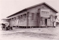 Causeway Hall just after construction. It has props against the walls. There is a car parked to the left hand side and two unidentified men near the entrance of the hall.