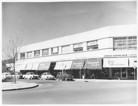 Corner of Garema Place and Alinga Street showing Roger Bartholomew's and Debenham's fashion store in Civic. There are several cars parked along the street in Garema Place.