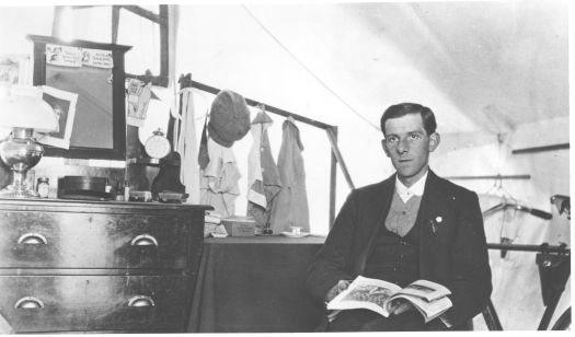 Man sitting with a book, living in a tent, surrounded by furniture.