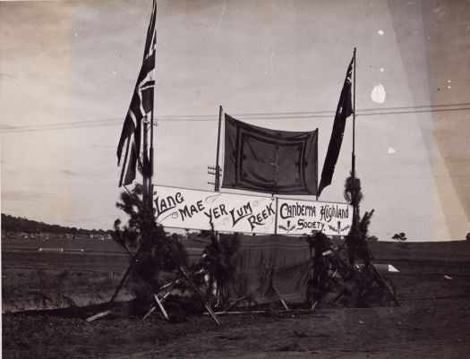 Sign reads 'Lang Mac Yer Lum Reek' 'Canberra Highland Society'. Sign is surmounted by a Scottish flag, with Union Jack, Australian flags mounted on each side. Photograph possibly taken adjacent to an aerodrome.