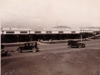 Street in Eastlake Shopping Centre (now Kingston) showing three parked cars and shops - The Federal Newsagency, S.O. Taylor Ladies and Gents Outfitters and the Southern Cross Dining and Supper Rooms.