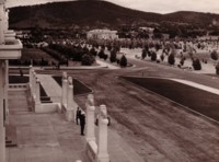 Shows a policeman standing at the top of the steps to old Parliament House and an unidentified man walking up the steps. There is a motor bike parked at the foot of the steps. The view is to the north west as Black Mountain, Albert Hall and Acton offices can be seen. Newly planted trees and parks are in the foreground.