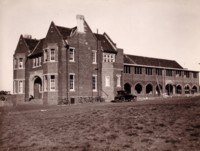 Photo taken from the left front side of the building. It shows a car and a number of bicycles leaning against a wall. An unknown person is sitting on the steps.