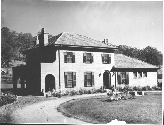 A front view of 'Roanoke', the home of Sir Robert Garran in Red Hill. It shows a circular driveway and flowers. Part of the house is two stories.