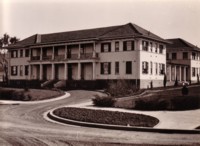 A view from the front left hand side of the building, with a curved driveway to the front steps, some small shrubs and a garden bed.