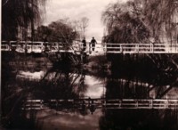 Two men standing on a footbridge over the Molonglo River. Their reflections can be seen as well as a number of willow trees.
