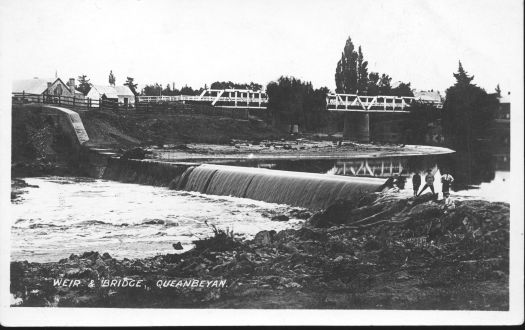 Shows bridge over the Queanbeyan River in the background. In the foreground four boys are watching the water pour over the weir wall.