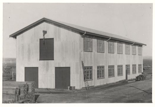 A two storey building with timber stacked outside the building. The building was near the Power House.
