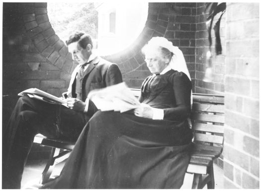 Robert Garran and his mother Mary, sitting on a bench in a brick porch and reading newspapers.