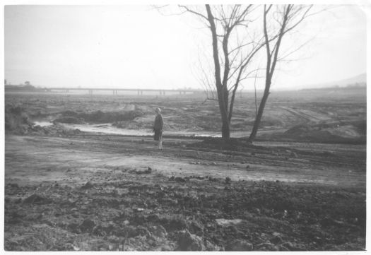 Photo shows Agnes Benson standing where East Basin in Lake Burley Griffin now is. Taken from in front of the Power House. Kings Avenue bridge is in the background and in between are earthworks associated with the lake.