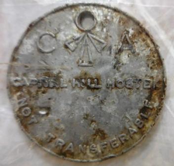 Metal disc, 35mm radius, with "Capital Hill Hostel, Not Transferable" on the face and "If Found put in Post Box, 22296, Department of Works and Housing CANBERRA ACT" on verso. A chain hole is at the top of the disc. Damaged but readable.