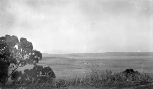 Undescribed view from a hill. A group of buildings are in the middle with hills in the distance.