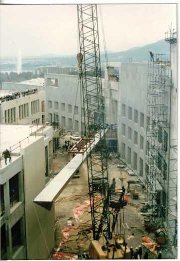 Crane raising the leg of the flagpole on the Senate side watched by a crowd of workers. The building is largely complete.