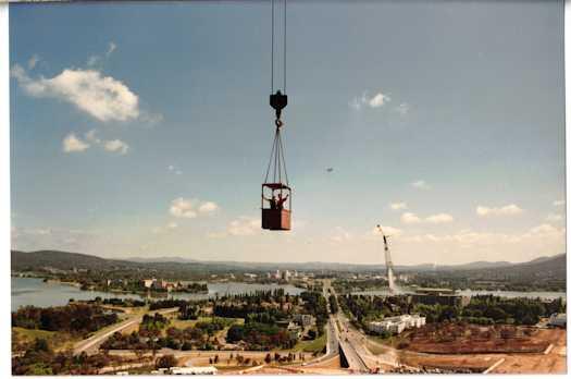 Two men (one is Alf Chambers) in the dog box, high in the air, moving around the site by crane. Royal Canberra Hospital is in the distance.