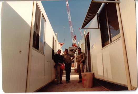 Three workmen (Alfred Chambers, Bob Taylor and unnamed) standing between worker's cribs. A crane is in the distance.