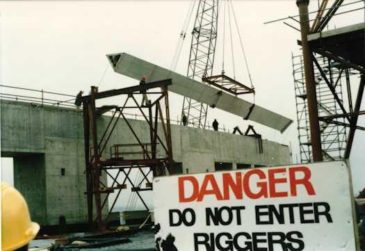 Straight section of the flag pole being raised by a crane. Sign says "Danger Do Not Enter Riggers".